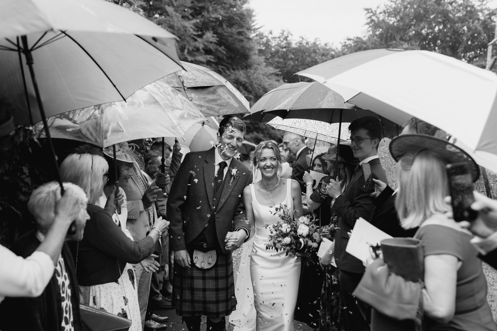 The bride and groom are greeted by a confetti tunnel in the rain