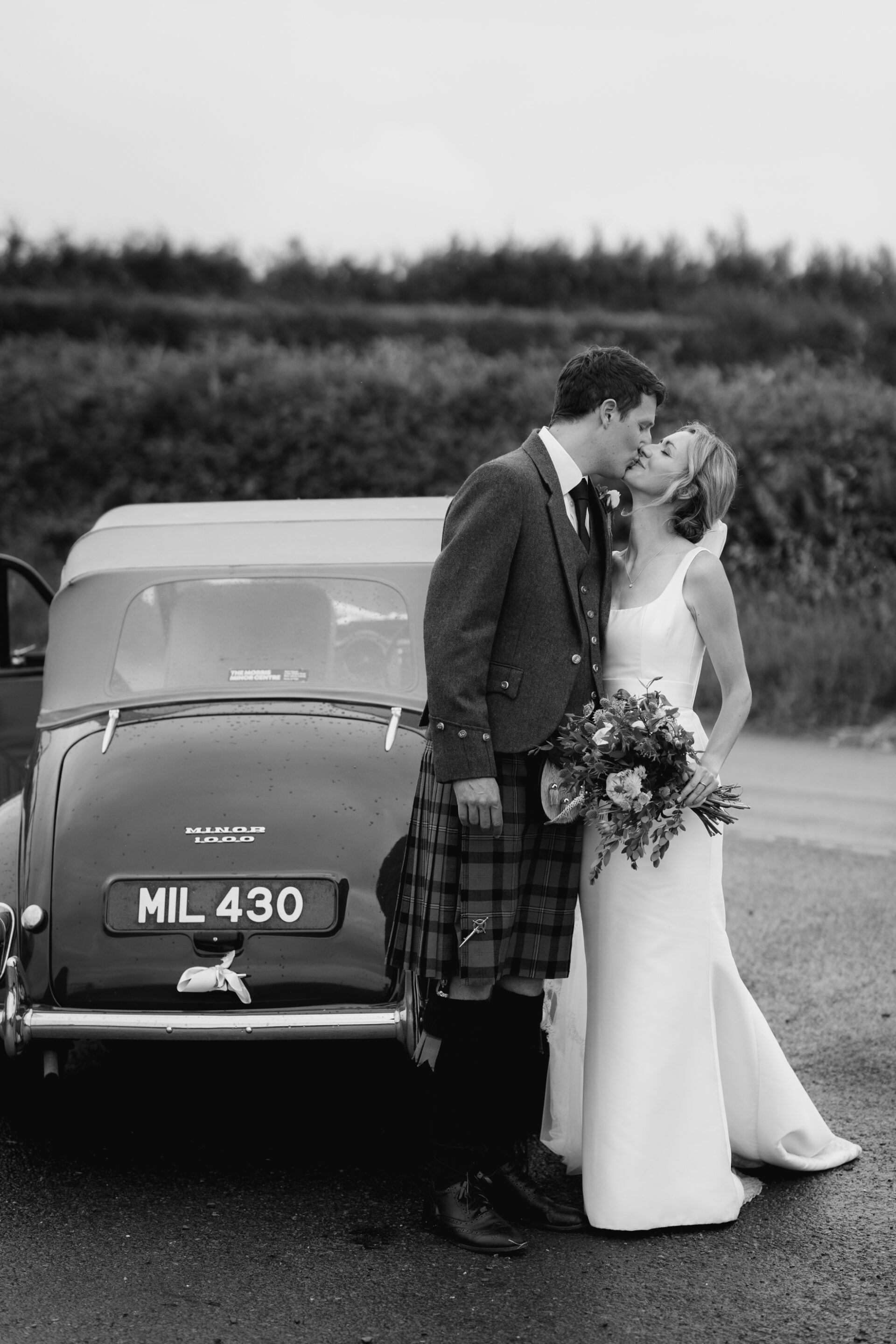 The bride and groom share a kiss next to their classic wedding car