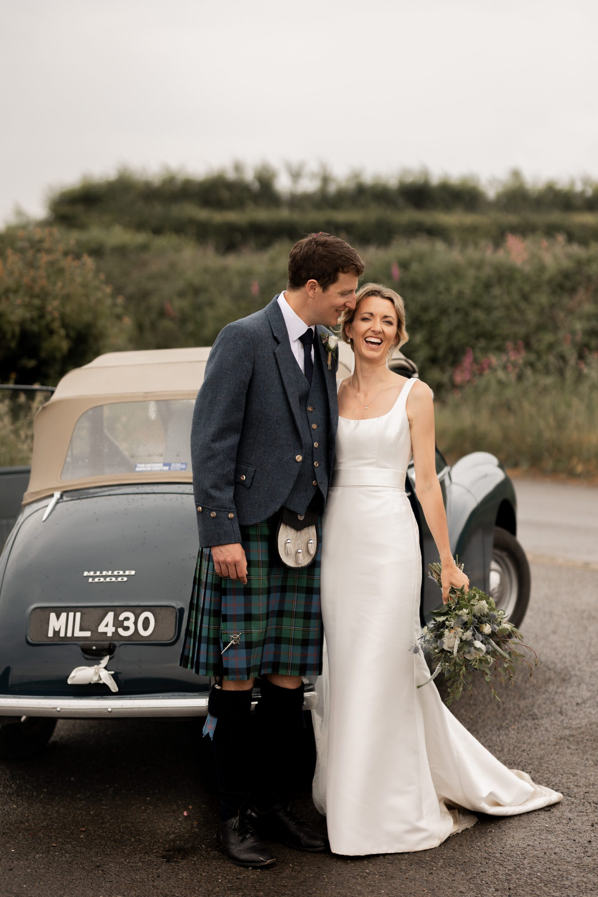 The bride and groom pose in front of their vintage wedding car in Devon