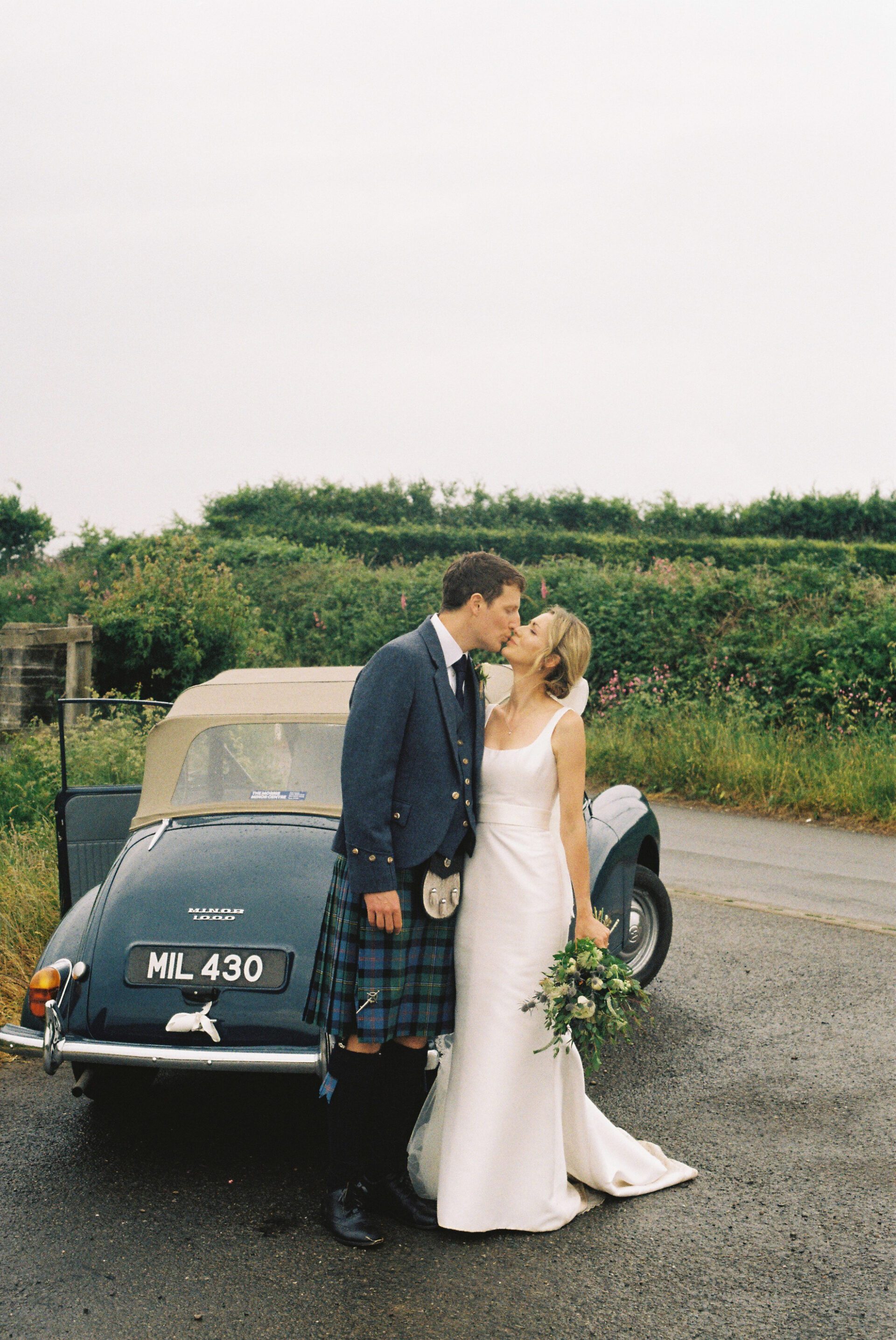 The bride and groom share a kiss in front of their vintage wedding car, captured on 35mm film