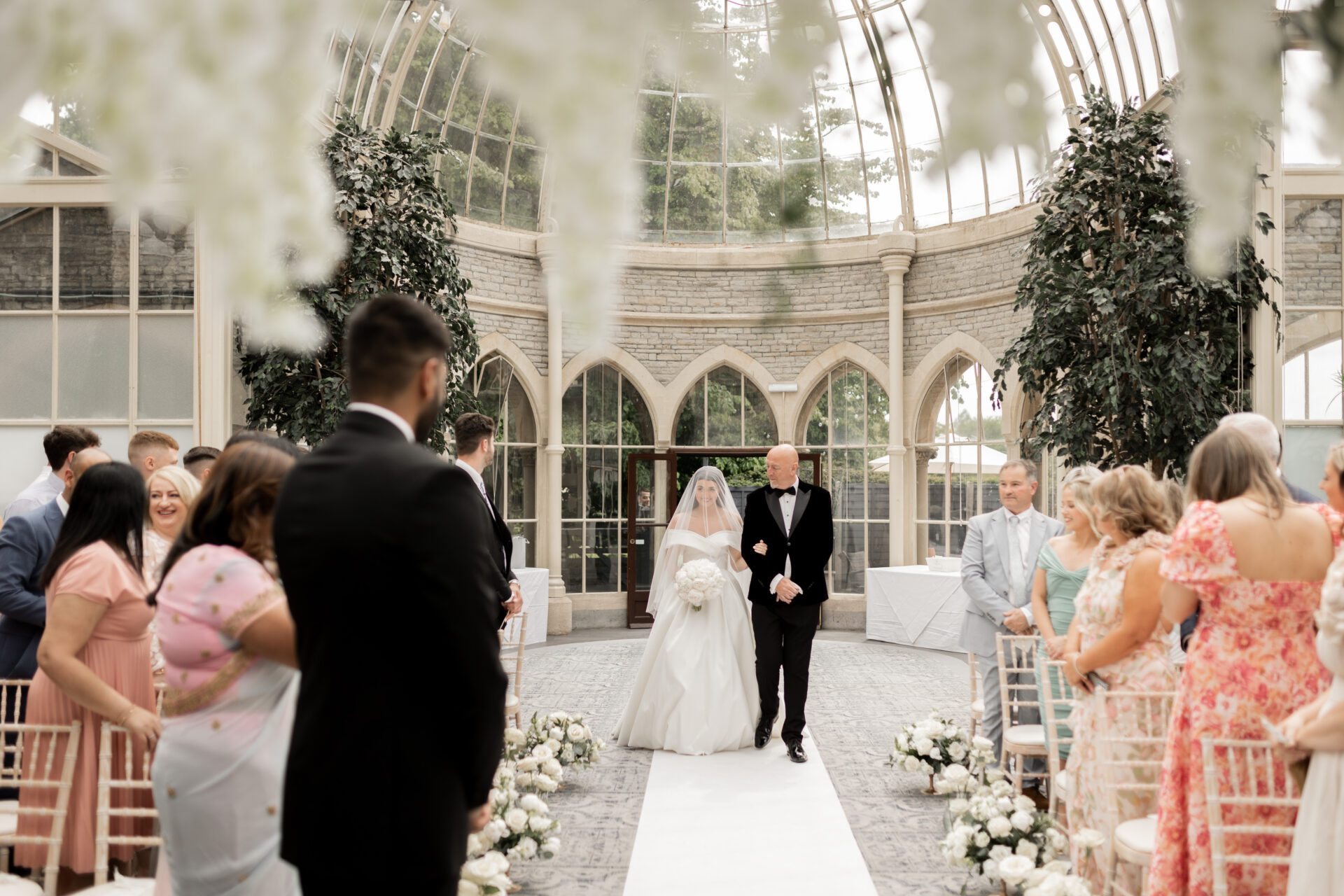 The bride and her father make their way down the aisle for the wedding ceremony in the Orangery at Tortworth Court