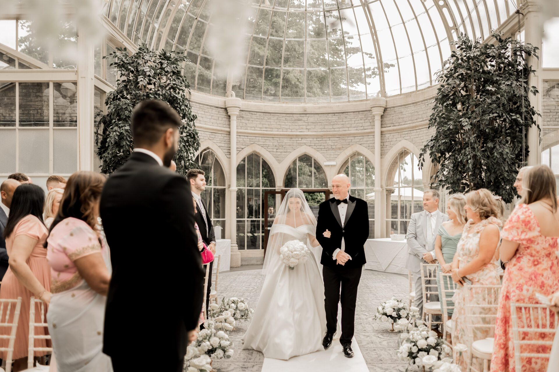 The bride and her father make their way down the aisle for the wedding ceremony in the Orangery at Tortworth Court